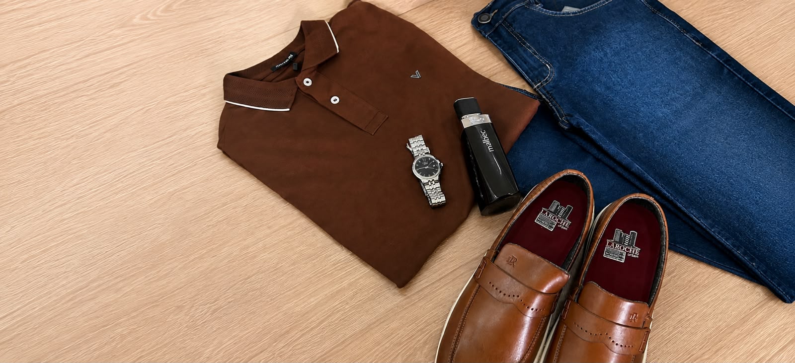 Camisa polo marrom, calça jeans, relógio, perfume e sapatos masculinos sobre mesa de madeira